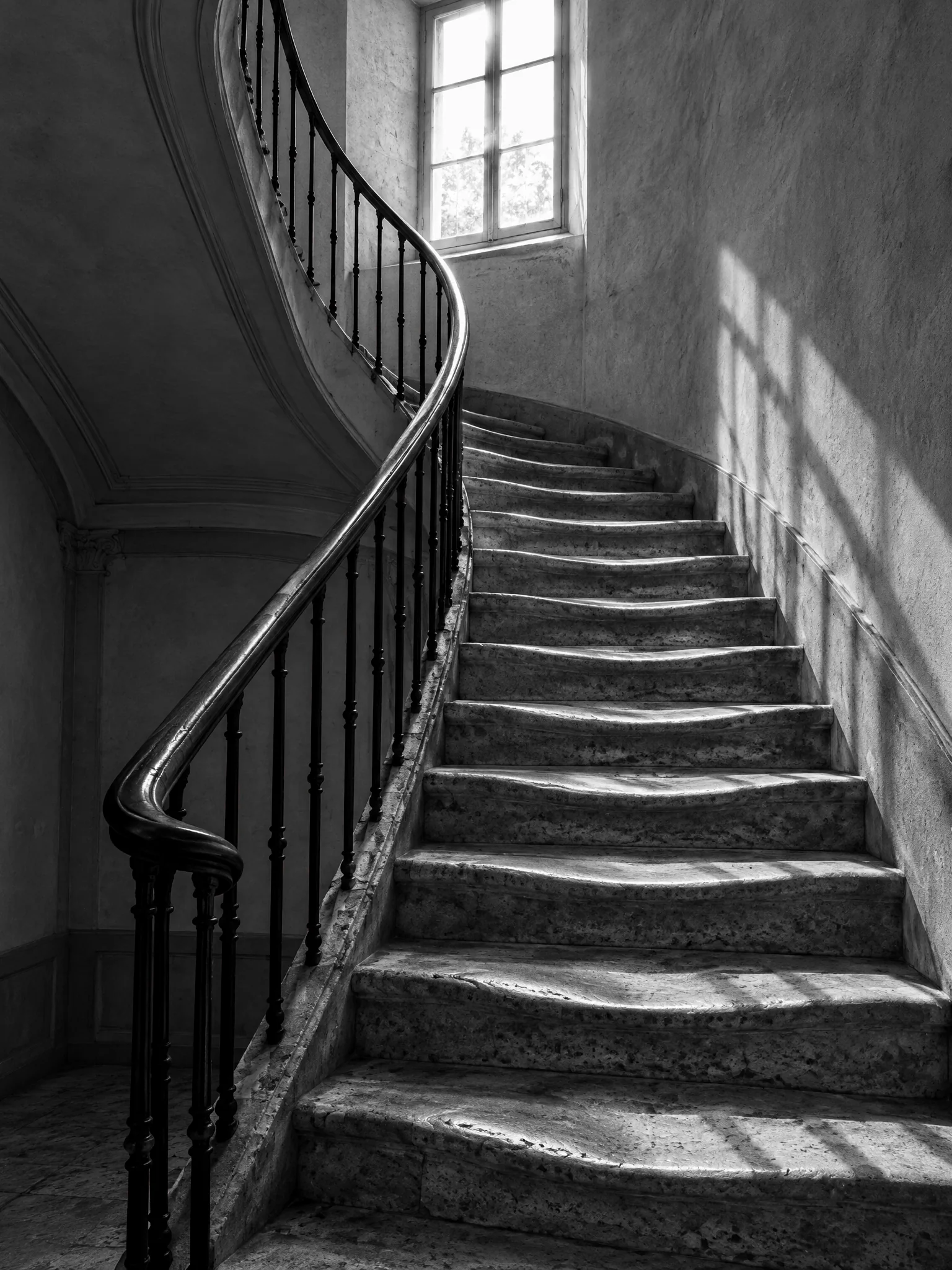 Escalera de piedra desgastada con una barandilla de hierro curva, iluminada por una ventana alta.